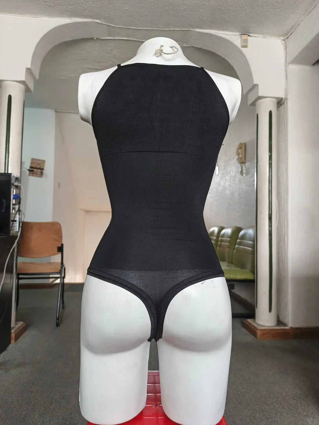 Maniquí femenino con body negro ajustado tipo tanga en sala con sillas y columnas